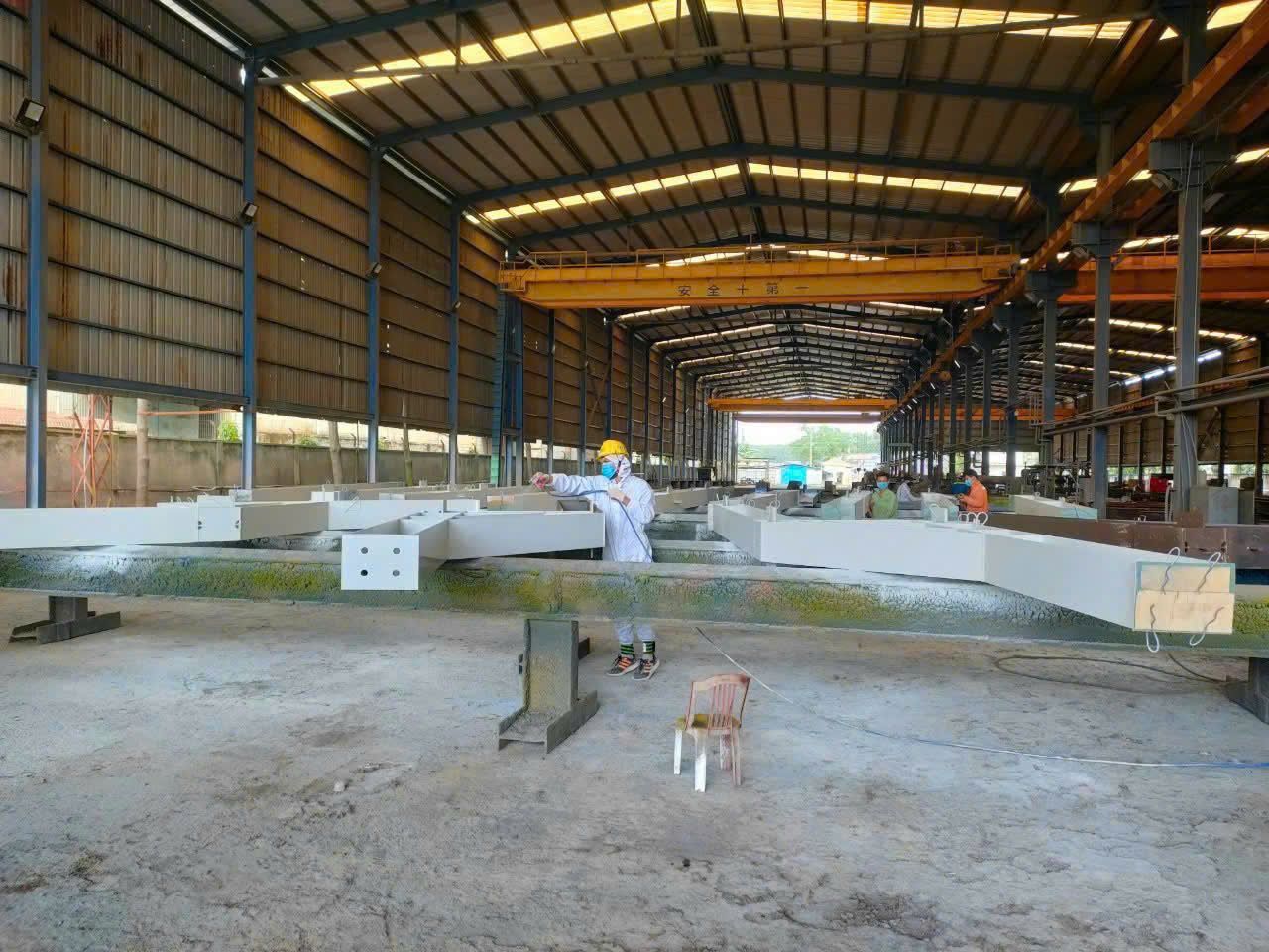 Công nhân mặc đồ bảo hộ toàn thân phun sơn dầm thép tại xưởng gia công có cầu trục