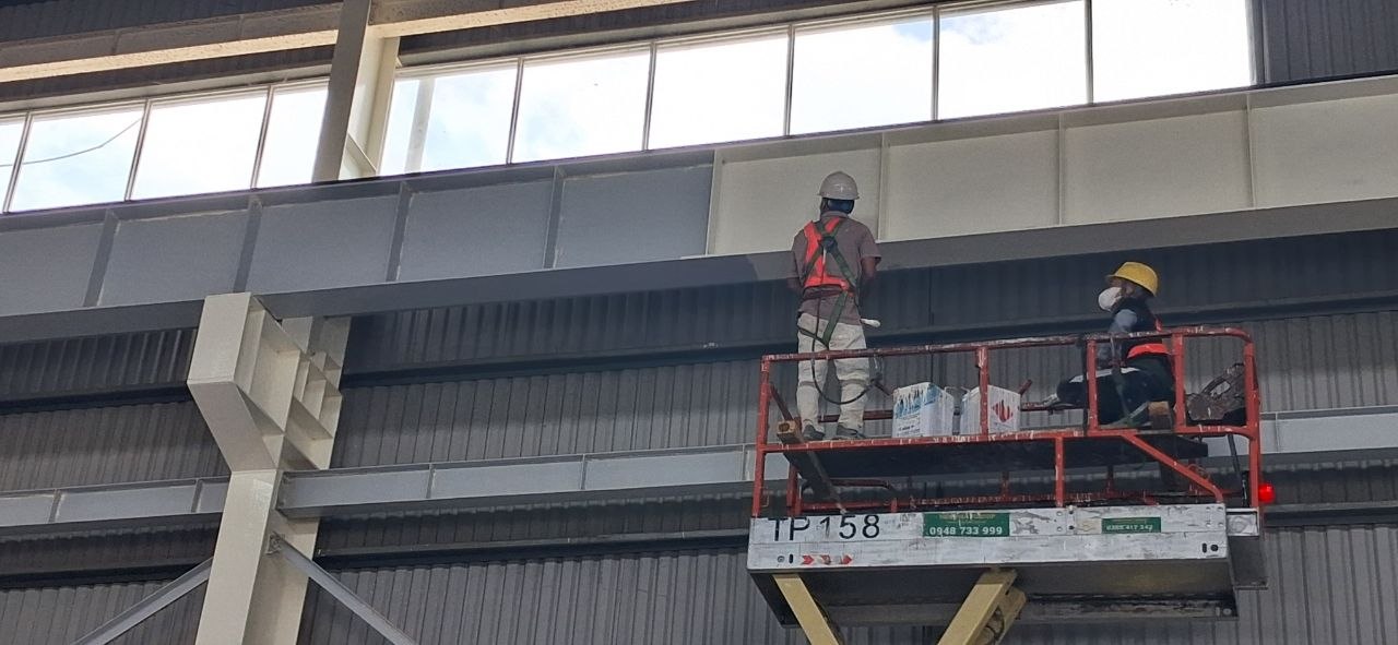 Hai công nhân thi công sơn kết cấu thép dầm nhà xưởng trên xe nâng người, có dây an toàn và bảo hộ đầy đủ
