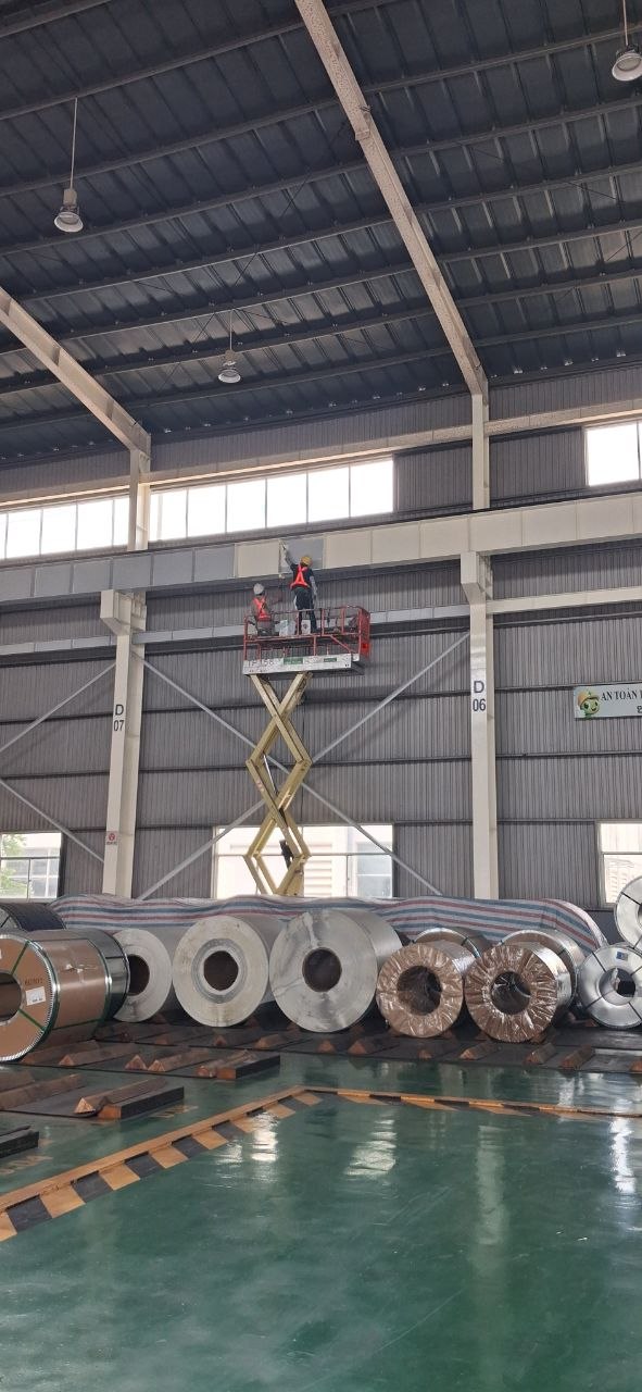 Toàn cảnh thi công sơn kết cấu thép trong nhà xưởng đang hoạt động, xe nâng người vươn cao, hàng hóa phía dưới